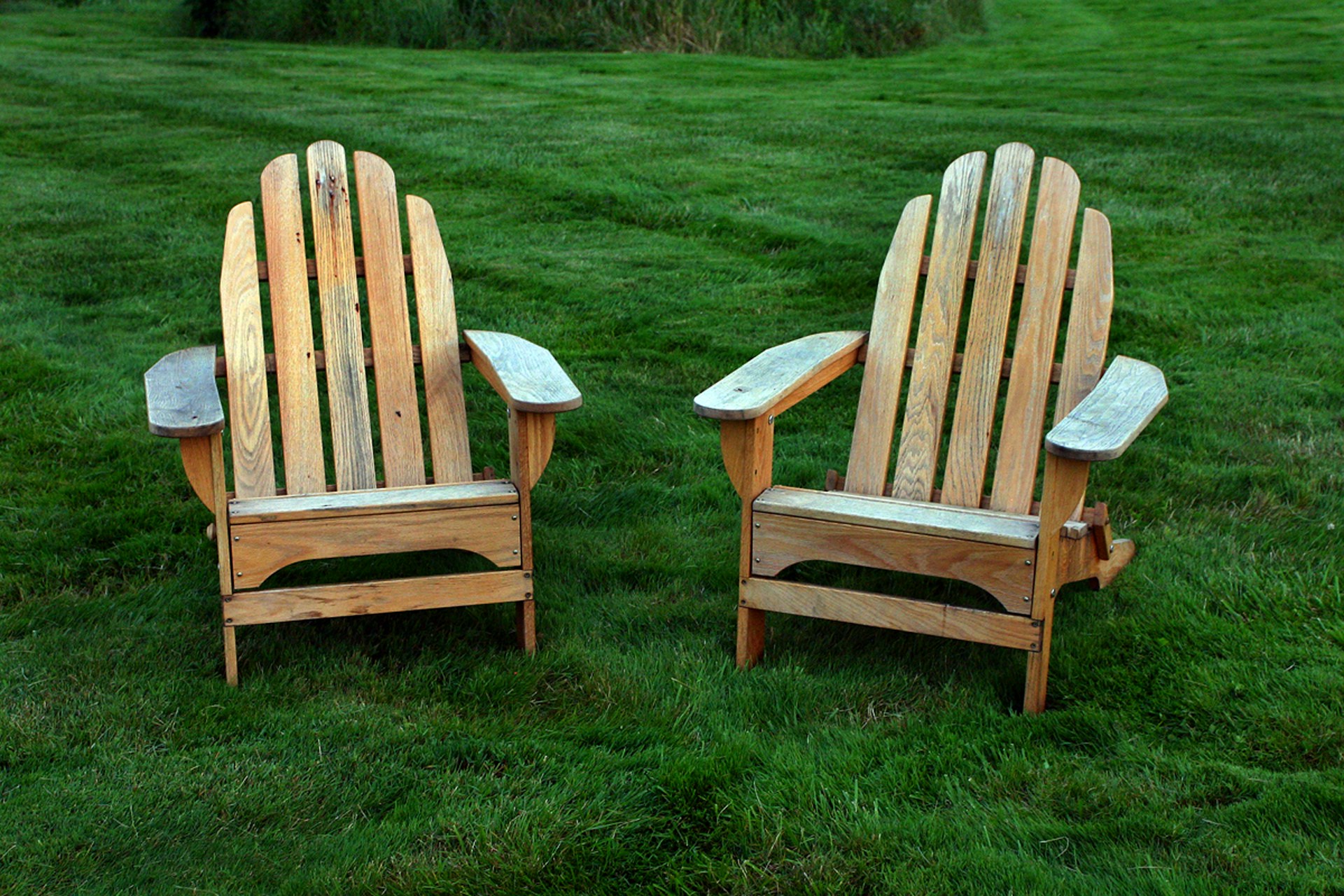 Two light colored wooden adirondack chairs on green grass