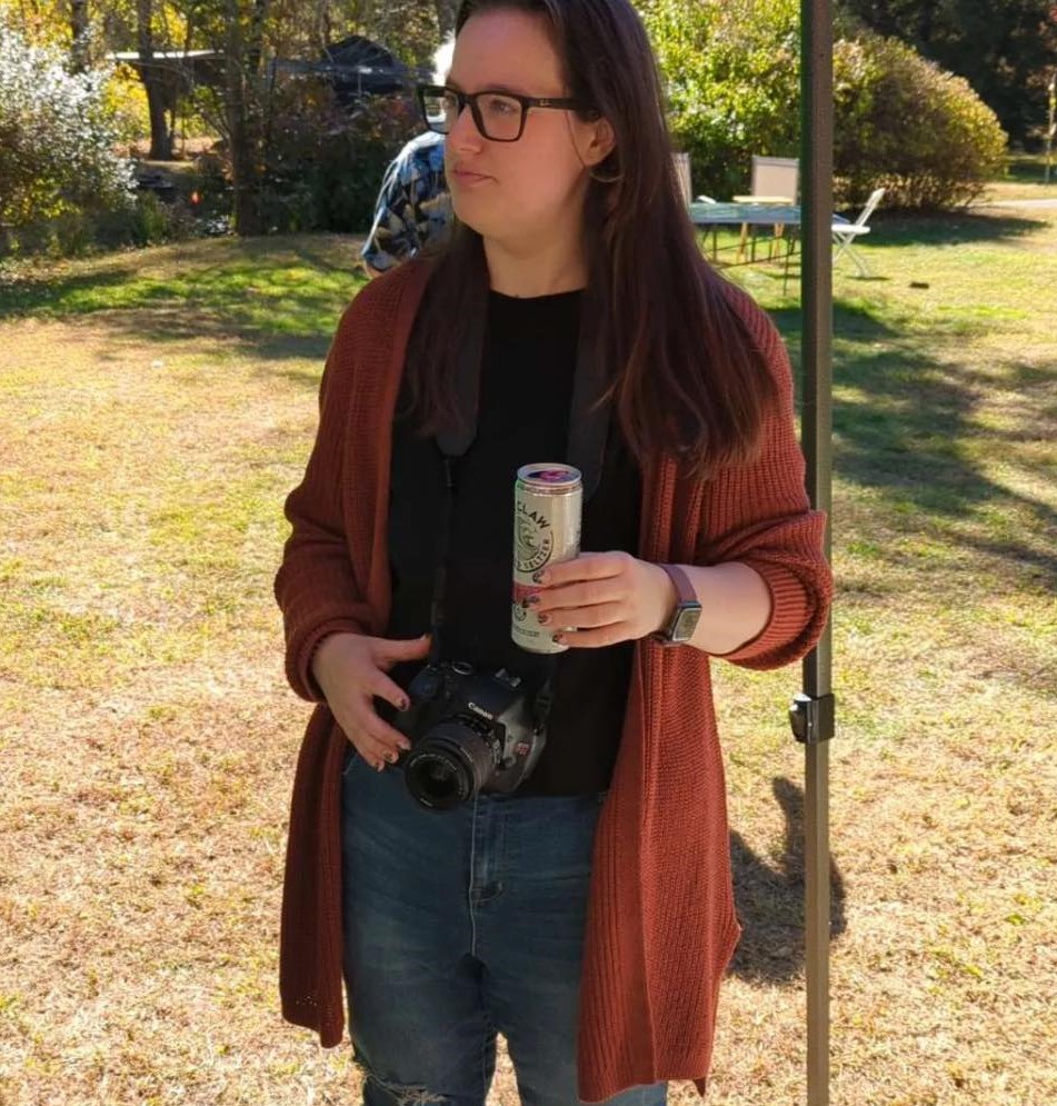 Picture of brunette woman with black shirt and orange cardigan and jeans. Holding a white claw can with camera around neck, looking to the left